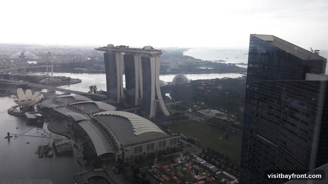 An aerial view of the Singapore skyline on a hazy day. Prominent landmarks include the Marina Bay Sands hotel with its distinctive three towers and rooftop infinity pool, the ArtScience Museum with its lotus-like design, and the domes of Gardens by the Bay. In the foreground, a modern glass skyscraper reflects the sky and surrounding buildings. The image also shows a body of water with boats, a Ferris wheel, and a distant coastline.