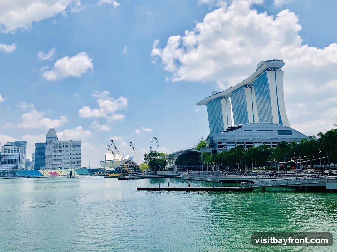 A wide shot of the Marina Bay Sands hotel and its distinctive three-tower design, connected by a rooftop infinity pool. The Singapore skyline is visible in the background, with other modern skyscrapers and a Ferris wheel. The foreground shows a body of water, likely Marina Bay, with a pier and some boats. The sky is blue with scattered white clouds.