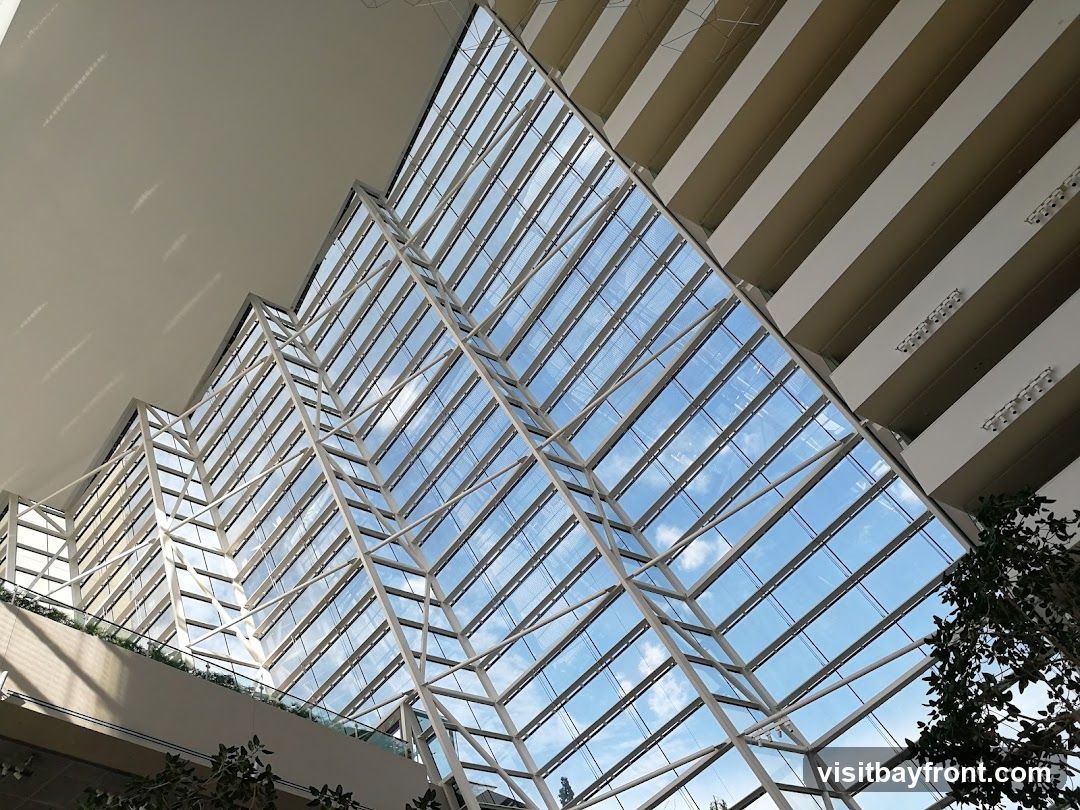 An interior view of a modern building atrium, looking upwards towards a large glass ceiling. The ceiling is supported by a white geometric metal framework, creating a striking visual pattern. Sunlight streams through the glass, illuminating the interior and casting shadows. The upper levels of the building, with their horizontal beige accents and lighting fixtures, are visible along the sides. Some greenery from indoor plants can be seen in the lower corners.