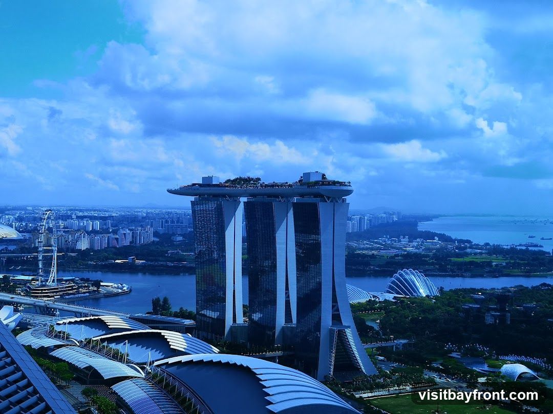 An aerial view of the Marina Bay Sands hotel in Singapore, with its iconic three towers and rooftop infinity pool. The surrounding cityscape includes the Singapore Flyer Ferris wheel, Gardens by the Bay, and other urban developments, all under a cloudy blue sky.