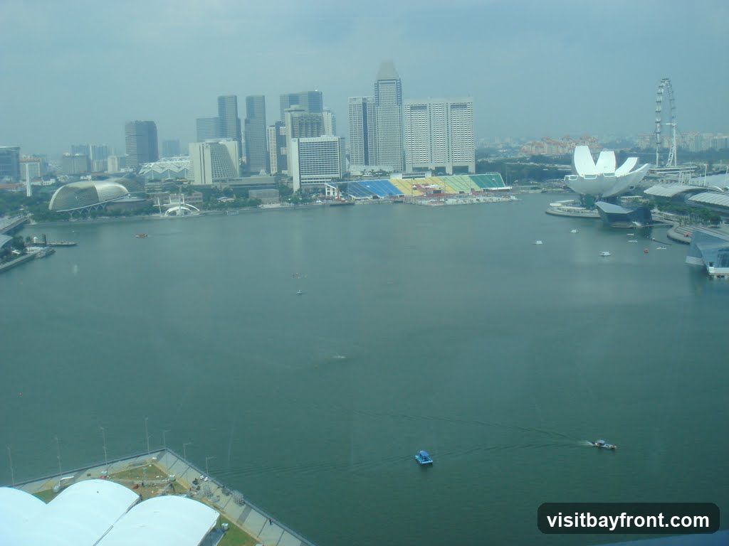 An aerial view of a city skyline with a large body of water in the foreground. Several modern buildings, including skyscrapers and distinctive architectural structures, are visible along the waterfront. A Ferris wheel and a unique lotus-shaped building are prominent landmarks. The water is dotted with small boats and a larger vessel. The sky is overcast, giving the scene a muted tone.