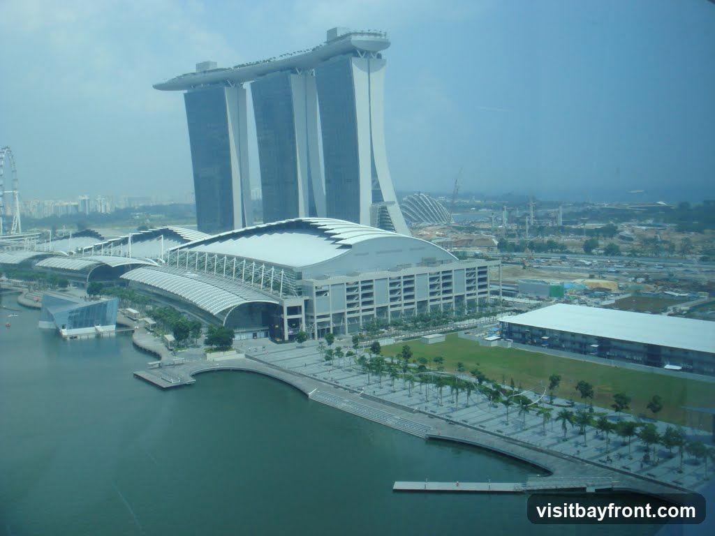 An aerial view of the Marina Bay Sands complex in Singapore, featuring the iconic three towers topped with a ship-like structure. In the foreground, a large convention center with a distinctive roof design is visible, situated next to a body of water. The surrounding area shows ongoing construction and urban development, with cranes and various buildings under construction. A Ferris wheel can be seen in the distance on the left side of the image. The sky is hazy and overcast.