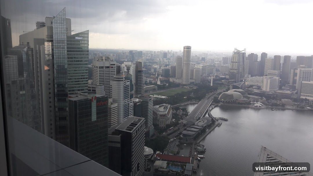 A panoramic view of a dense cityscape dominated by numerous skyscrapers of varying heights and architectural styles. A body of water, likely a river or bay, runs through the scene, with bridges and waterfront structures visible. The sky is overcast, casting a diffused light over the urban landscape. Several buildings have visible signage, including 'Keppel' and 'NTUC'.