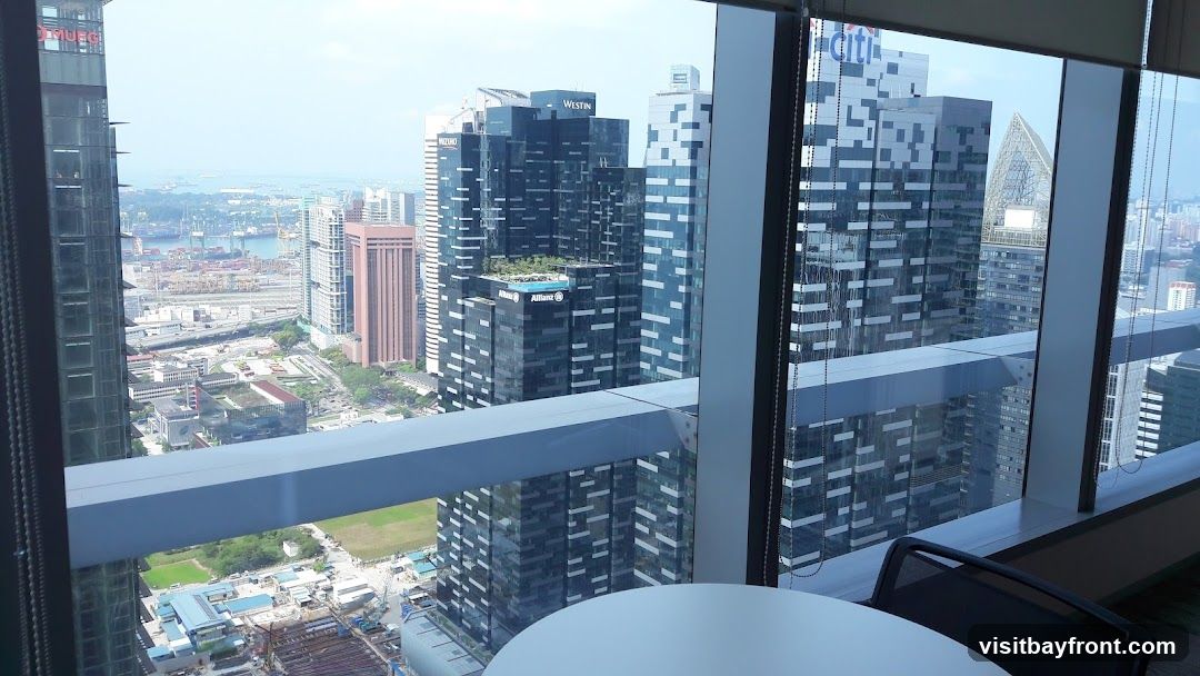 A panoramic view of a bustling city skyline, seen through a large office window. In the foreground, a white circular table and a black office chair are partially visible. The mid-ground features a dense collection of modern skyscrapers, some with distinctive architectural designs and visible signage such as 'Westin' and 'Allianz'. Beyond the buildings, a large port with numerous ships and shipping containers is visible, stretching towards the horizon under a hazy sky. The overall impression is one of a vibrant, active urban environment from a high vantage point.