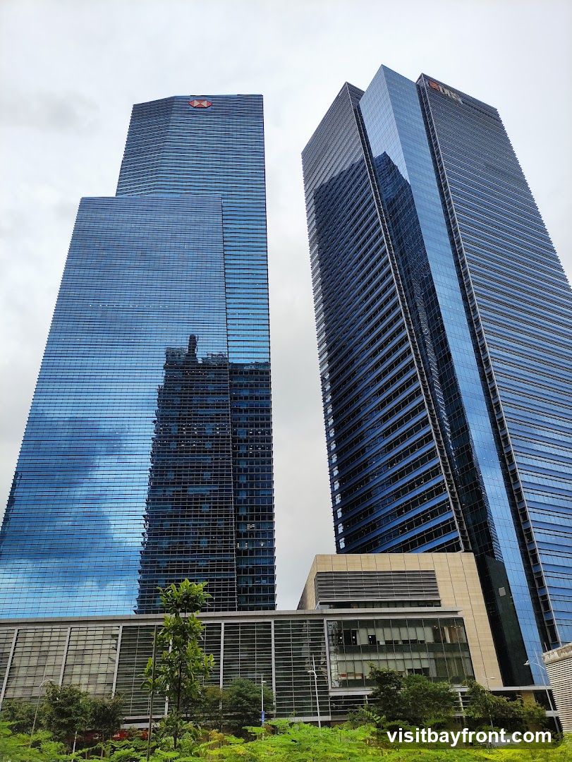 A low-angle shot of two modern skyscrapers with blue glass facades against a cloudy sky. The skyscraper on the left has a red circular logo near its top. The skyscraper on the right has a red logo near its top. Green trees and foliage are visible at the base of the buildings.