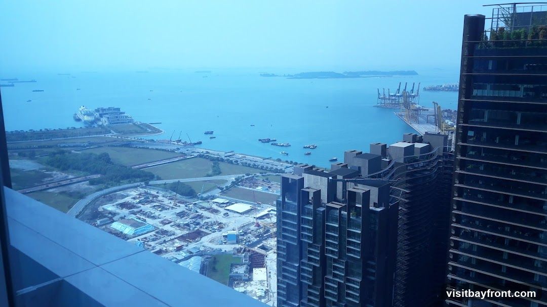 An aerial view of a city with a large harbor. In the foreground, modern skyscrapers dominate the right side of the frame. To the left, a sprawling urban landscape with construction sites and roads is visible. The harbor is filled with various ships, including a large cruise ship docked at a pier. In the distance, more ships and islands can be seen under a hazy sky.