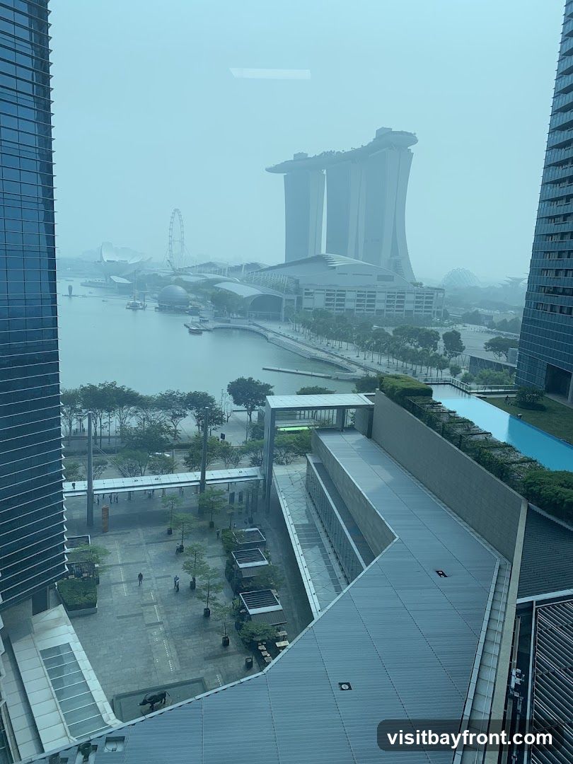 A hazy view of a cityscape, likely Singapore, featuring the iconic Marina Bay Sands hotel and other prominent buildings. The image captures a waterfront area with a Ferris wheel in the distance. In the foreground, modern architecture with glass facades and outdoor seating areas are visible. The overall atmosphere is subdued due to the haze.