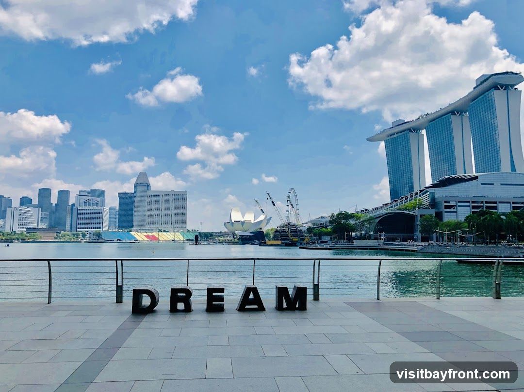 A scenic view of the Singapore skyline on a bright, cloudy day. In the foreground, large black letters spell out "DREAM" on a paved walkway. The water in the bay reflects the blue sky and clouds. In the background, various skyscrapers and iconic buildings, including the Marina Bay Sands, are visible. A Ferris wheel and construction cranes are also present, suggesting ongoing development or attractions. The overall scene captures a modern urban landscape with a touch of inspirational messaging.