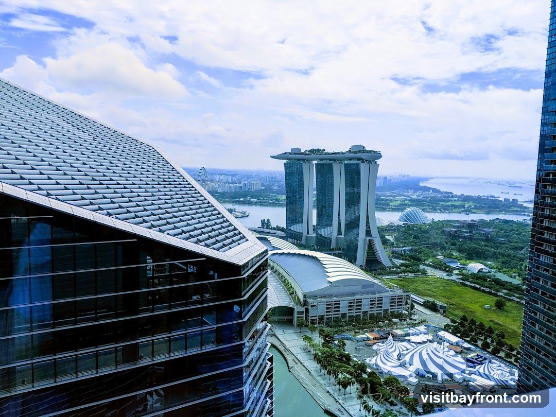 A panoramic view of a cityscape, dominated by the iconic Marina Bay Sands hotel in Singapore. In the foreground, a modern building with a distinctive angled glass roof is visible. To the right, another tall skyscraper stands. In the middle ground, the Marina Bay Sands complex with its three towers and rooftop infinity pool is prominent. Below it, the Gardens by the Bay with its Supertree Grove and conservatories can be seen. A large, striped tent structure is set up on the waterfront, suggesting an event or temporary venue. The sky is partly cloudy, with a bright blue expanse.