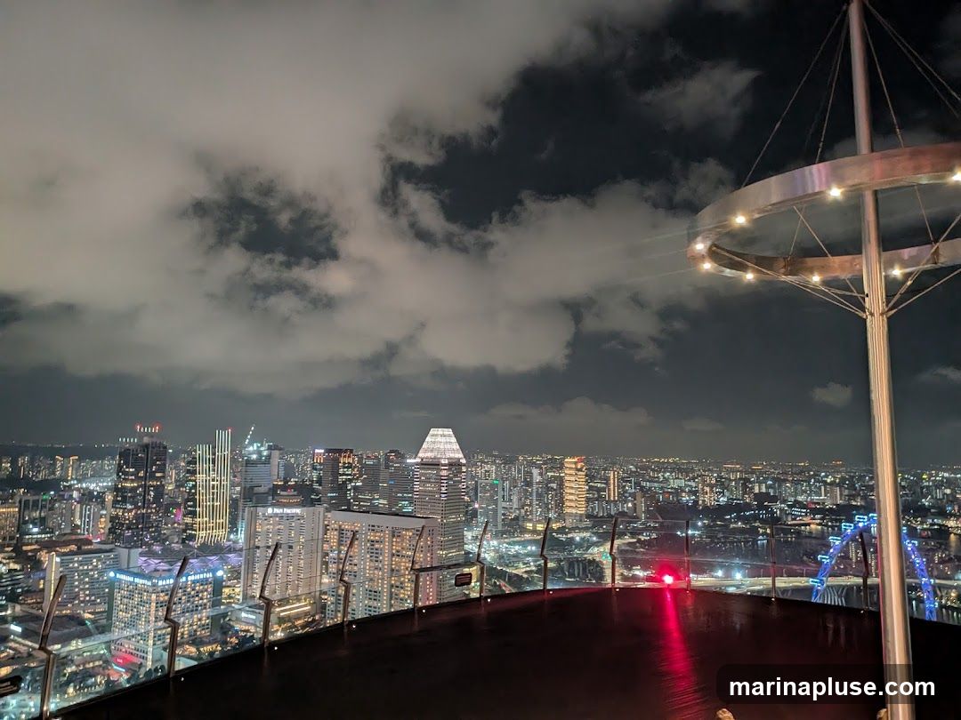 A panoramic view of a sprawling cityscape at night, seen from a high vantage point. The foreground features a dark, reflective surface, possibly a rooftop bar or observation deck, with a metallic structure adorned with lights. Beyond this, a dense collection of illuminated skyscrapers and buildings stretches into the distance. Clouds drift across the dark sky, partially obscuring the city lights. A large Ferris wheel is visible on the right side of the frame, glowing with blue lights.
