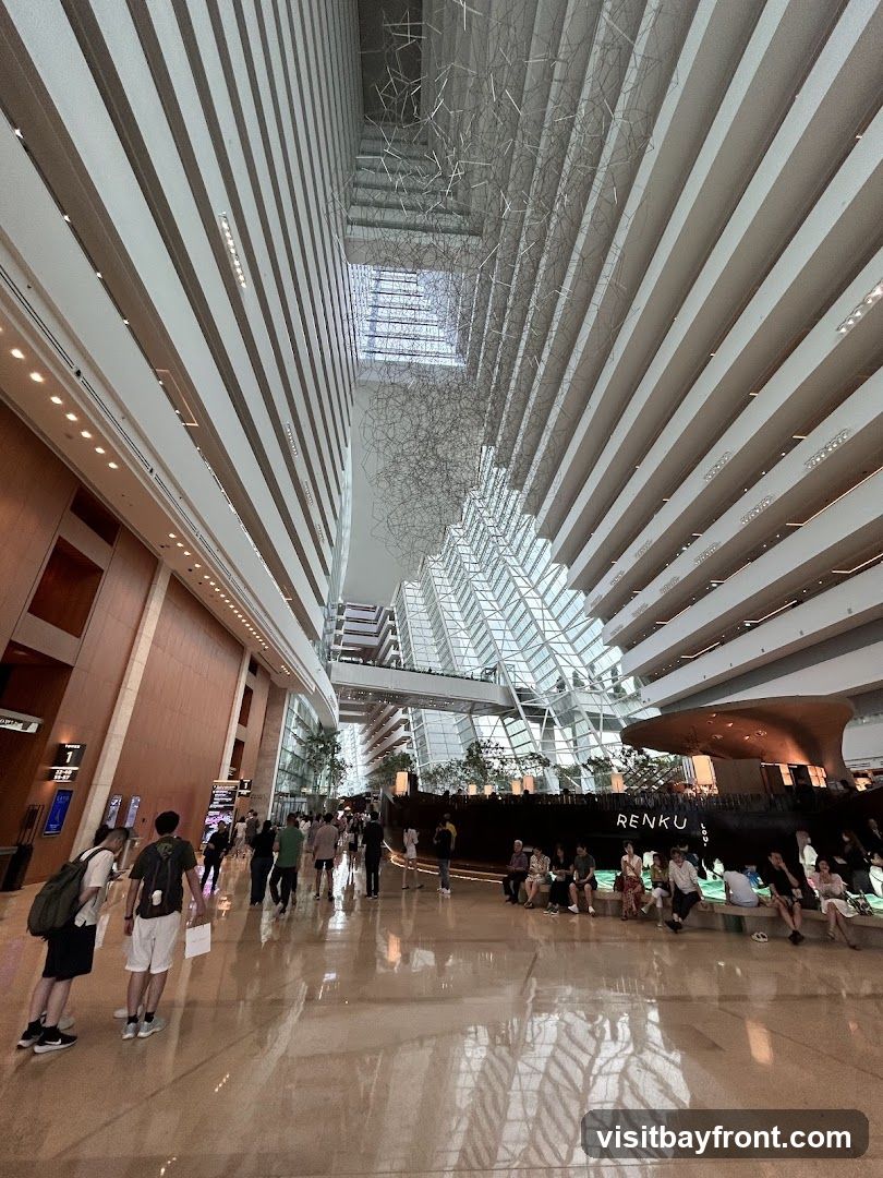 The image shows the interior of a vast atrium with a high ceiling. The architecture features multiple levels of balconies or walkways overlooking a central open space. A large, abstract metal sculpture hangs from the ceiling. People are seen walking and sitting in the atrium. In the background, there is a large, dark structure with the word "RENKU" visible, possibly a restaurant or bar. The floor is highly reflective, mirroring the architecture above.