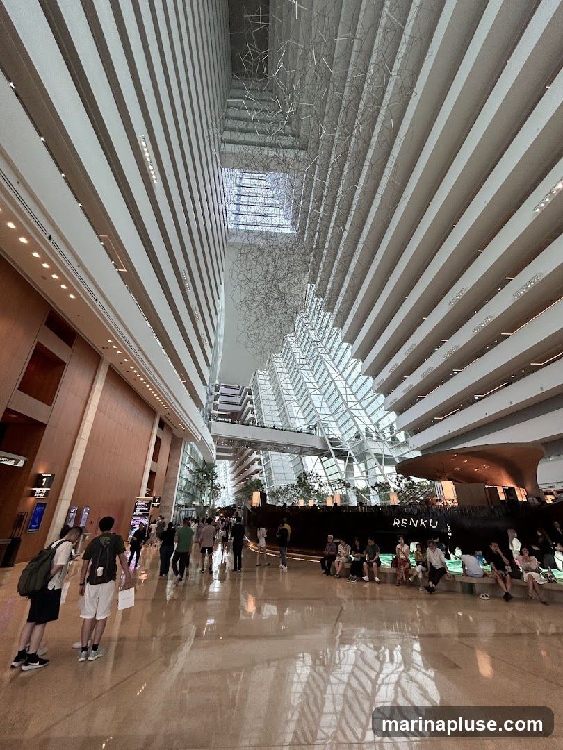 The image shows the interior of a vast atrium with a high ceiling. The architecture features multiple levels of balconies or walkways overlooking a central open space. A large, abstract metal sculpture hangs from the ceiling. People are seen walking and sitting in the atrium. In the background, there is a large, dark structure with the word "RENKU" visible, possibly a restaurant or bar. The floor is highly reflective, mirroring the architecture above.
