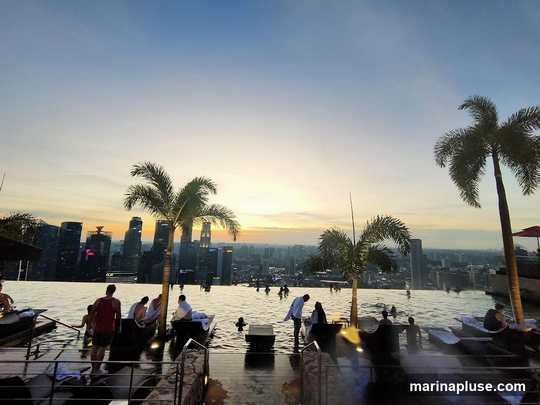 People relax in an infinity pool with a panoramic view of a city skyline at sunset. Palm trees are scattered around the pool area. The sky is a gradient of blue and orange, with the sun low on the horizon.