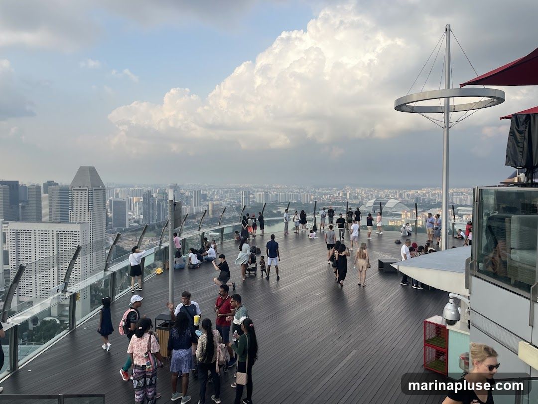 A wide shot of a crowded rooftop observation deck overlooking a sprawling cityscape under a cloudy sky. People are scattered across the wooden deck, some sitting, some standing, and some walking. In the foreground, a woman with blonde hair is walking towards the camera. The background shows numerous tall buildings and a hazy horizon. There are umbrellas and seating areas visible on the deck.