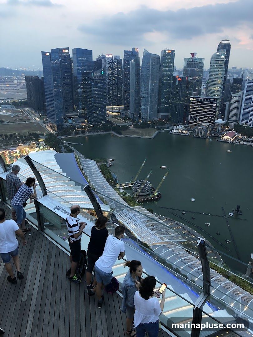 A group of people stand on a wooden deck, looking out at the Singapore skyline at dusk. The view includes numerous skyscrapers, the Marina Bay waterfront, and the distinctive architecture of the Marina Bay Sands hotel. The sky is cloudy, and the city lights are beginning to illuminate the scene.