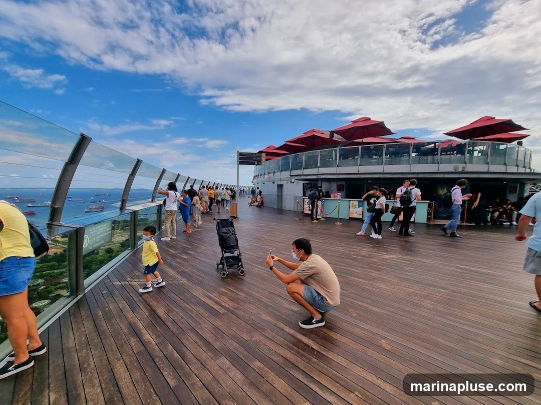 People are enjoying the view from a high vantage point, with a cityscape and ocean visible in the background. The foreground features a wooden deck with several individuals, including a man taking a photo and a child walking. In the mid-ground, there is a building with red umbrellas on its rooftop, suggesting an outdoor seating area for a restaurant or bar.