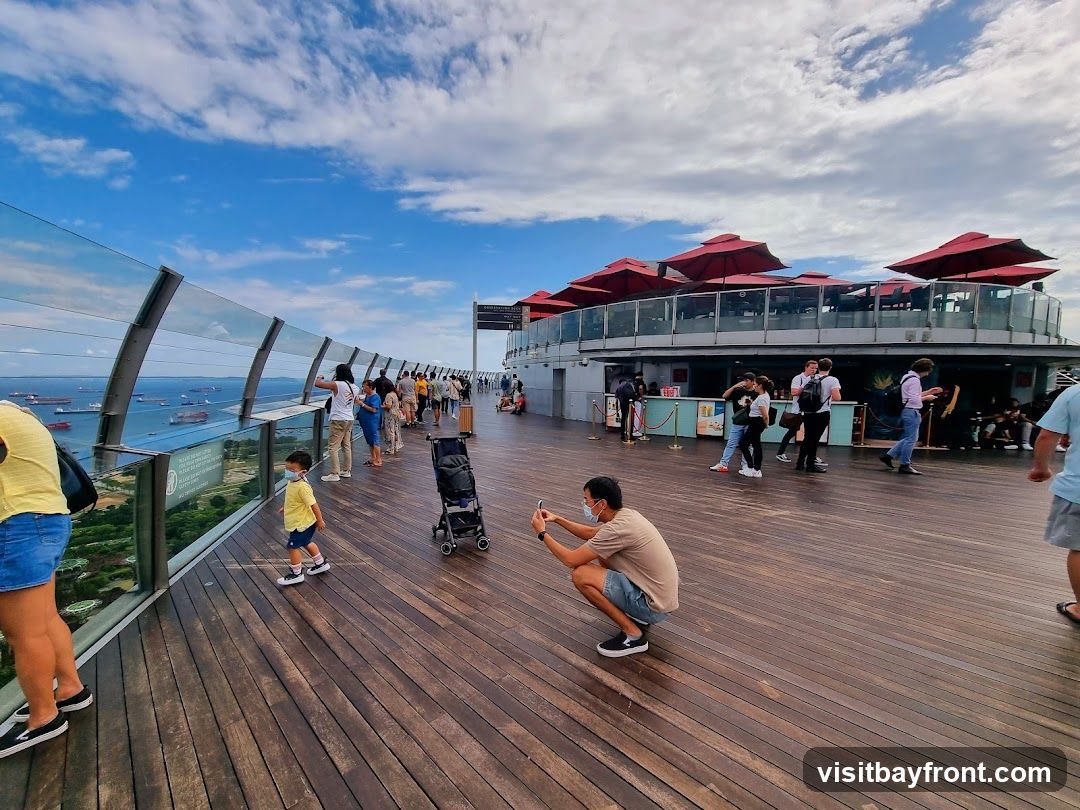 People are enjoying the view from a high vantage point, with a cityscape and ocean visible in the background. The foreground features a wooden deck with several individuals, including a man taking a photo and a child walking. In the mid-ground, there is a building with red umbrellas on its rooftop, suggesting an outdoor seating area for a restaurant or bar.