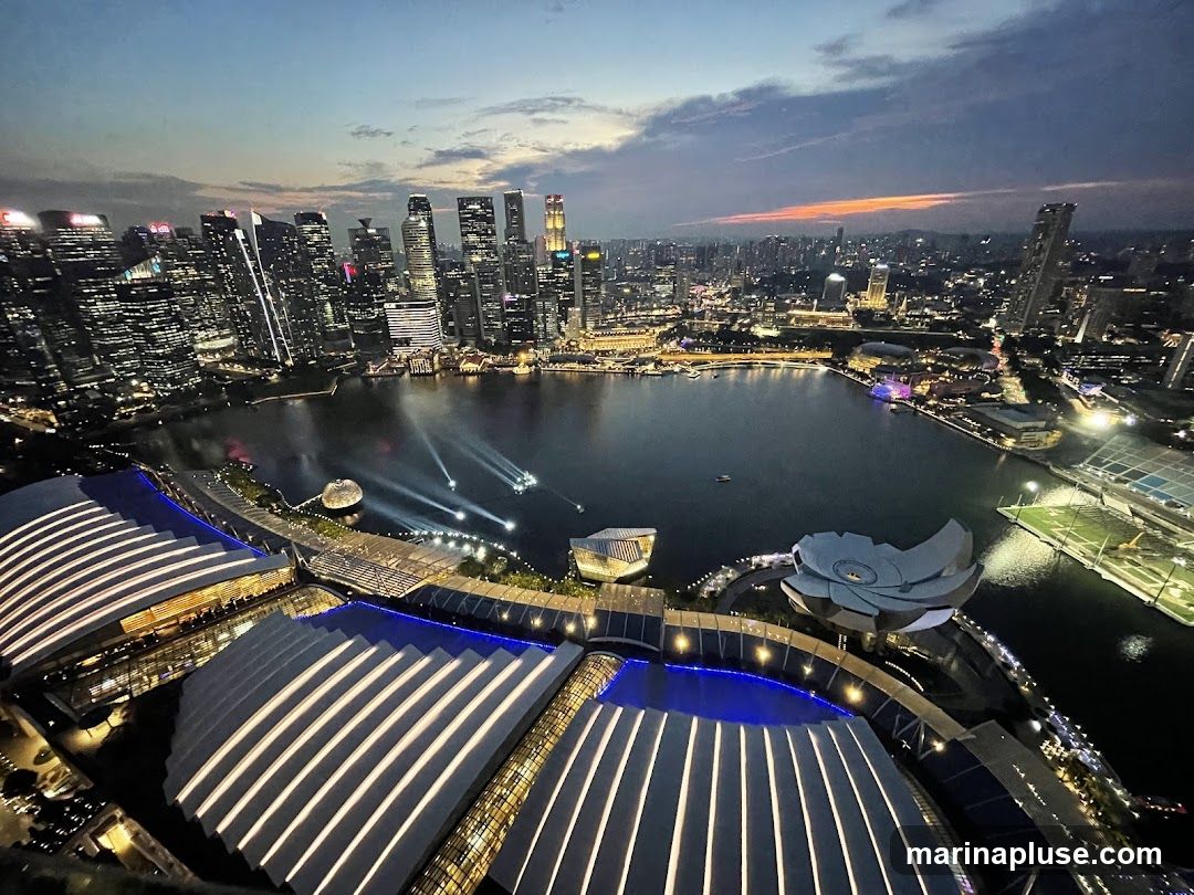 An aerial view of a modern cityscape at dusk, with a body of water in the foreground reflecting the lights of surrounding buildings. Several illuminated structures, possibly part of a park or entertainment complex, are visible in the lower portion of the image. In the distance, a dense cluster of skyscrapers forms the city skyline, with lights twinkling from many windows. The sky above is a gradient of deep blue and orange, indicating the setting sun. Beams of light are projected onto the water from a point on the shore.