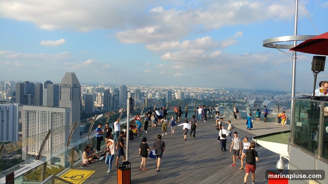 A panoramic view of a city skyline from a rooftop bar. Many people are gathered on the wooden deck, enjoying the view and socializing. A DJ booth with speakers is visible on the right, and a person is serving drinks. The sky is blue with scattered clouds.