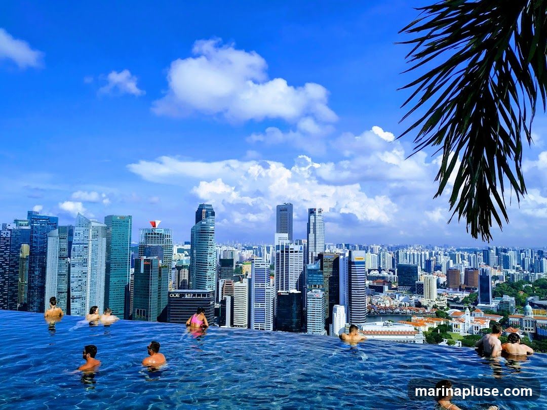 An infinity pool with several people relaxing in the water, set against a backdrop of the Singapore skyline. Tall skyscrapers dominate the view under a bright blue sky with scattered clouds. A palm frond hangs in the upper right corner, framing the scene.