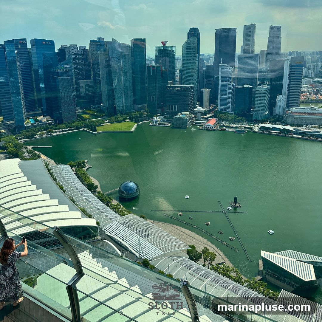 A high-angle shot captures the expansive Singapore skyline, featuring numerous modern skyscrapers, including buildings with visible logos like HSBC and UOB. The lush green expanse of a park is situated between the city and the shimmering green waters of Marina Bay. A distinctive glass dome structure is visible in the bay, along with various floating platforms and boats. In the foreground, a woman stands on a balcony, taking a photograph with her phone, overlooking the scenic view. The architecture of the building she is on, with its white, curved roof structures, is also prominent. A watermark with a van and the text 'SLOTH TRIP' is present on the lower right side of the image.
