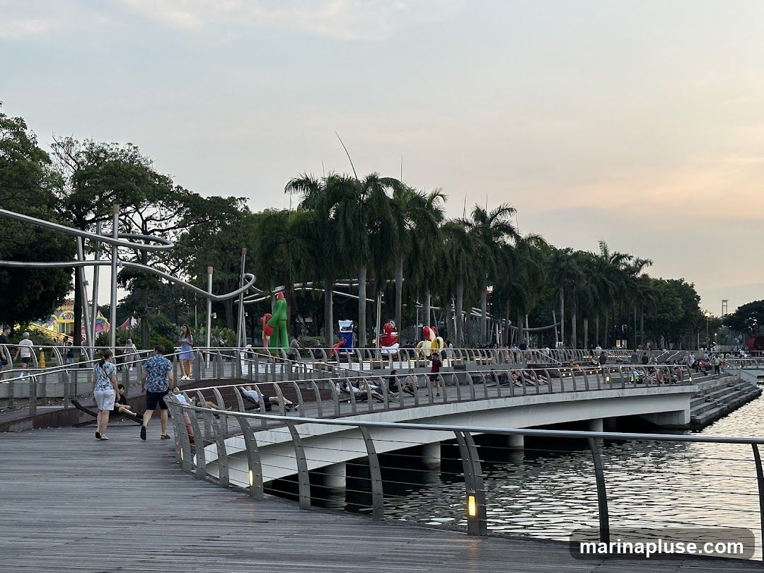 A wooden boardwalk curves along a body of water, lined with palm trees and people. In the background, there are various colorful sculptures and structures, including a tall green figure and red and yellow balloon-like figures. People are seen walking, sitting, and relaxing on the boardwalk and along the water's edge. The sky is a soft gradient of pale orange and blue, suggesting it is either sunrise or sunset.