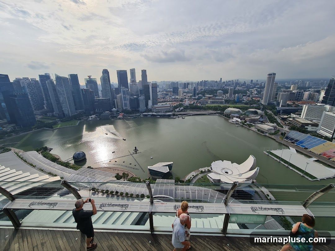 A wide-angle view of the Singapore skyline from a high vantage point, overlooking Marina Bay. The image captures numerous skyscrapers, a body of water with boats, and distinctive architectural structures. Several people are visible on a wooden deck in the foreground, some taking photographs. The sky is partly cloudy.