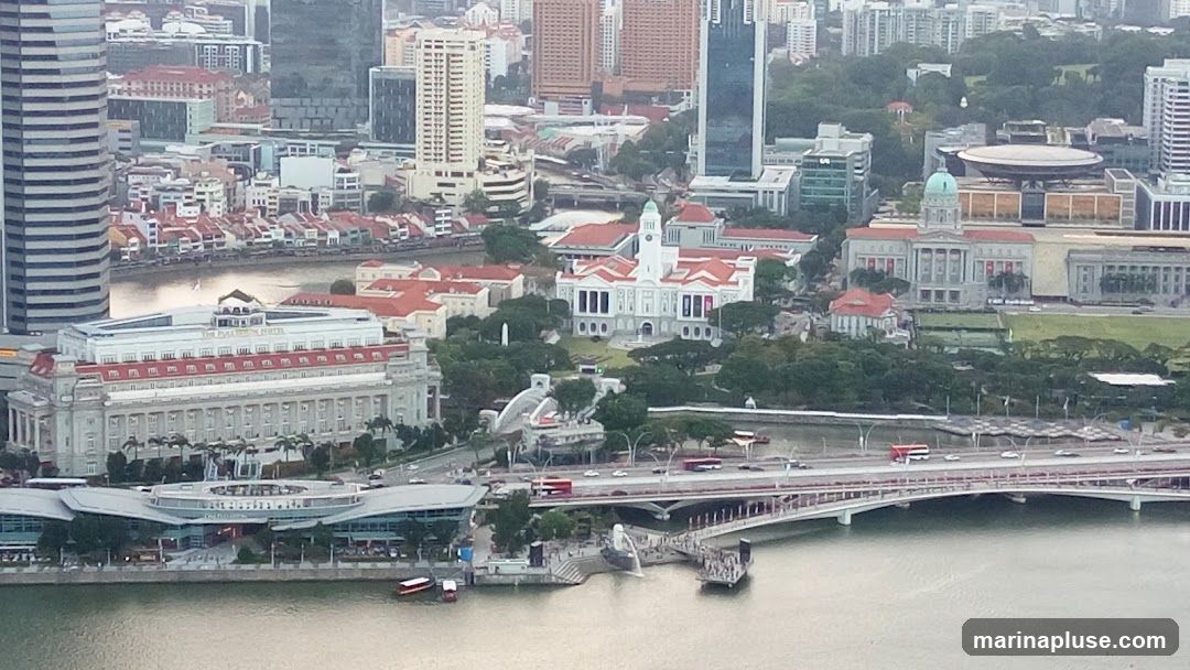 An aerial view of a city with a river running through it. Several bridges span the river, with traffic visible on them. Buildings of various architectural styles and heights are scattered throughout the cityscape, including a prominent white building with a red roof and a large, modern glass structure. Green spaces and trees are interspersed among the buildings. The overall scene depicts a bustling urban environment.