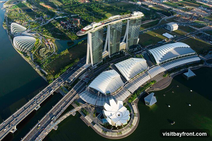 An aerial view of the iconic Marina Bay Sands hotel and the Gardens by the Bay in Singapore. The image captures the three towering hotel buildings connected by the SkyPark, as well as the distinctive Supertree Grove and the large conservatories of Gardens by the Bay. Several bridges and roads with traffic are visible, along with the surrounding urban landscape and water bodies.