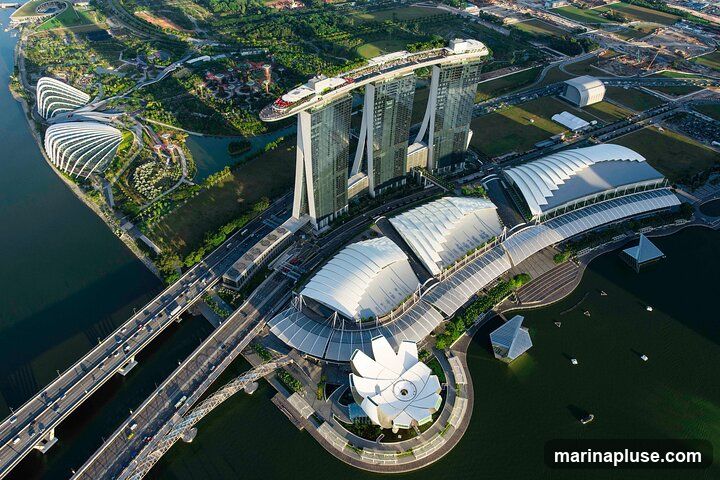 An aerial view of the iconic Marina Bay Sands hotel and the Gardens by the Bay in Singapore. The image captures the three towering hotel buildings connected by the SkyPark, as well as the distinctive Supertree Grove and the large conservatories of Gardens by the Bay. Several bridges and roads with traffic are visible, along with the surrounding urban landscape and water bodies.