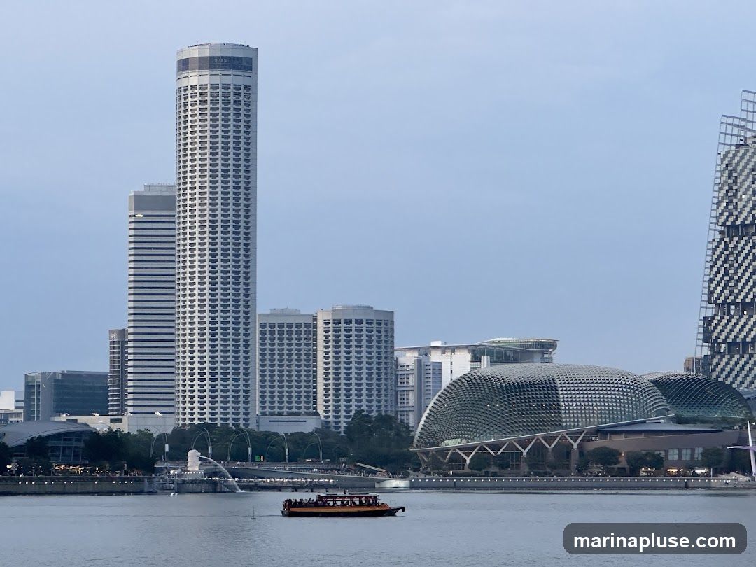A panoramic view of the Singapore skyline at dusk, featuring iconic landmarks such as the Marina Bay Sands hotel, the Esplanade - Theatres on the Bay, and the Fullerton Hotel. A boat is visible on the water in the foreground, and the Merlion statue can be seen in the distance. The sky is a soft blue, indicating the transition from day to night.