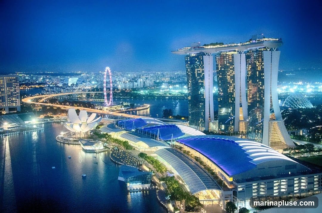 A panoramic view of Marina Bay Sands in Singapore at night. The iconic three-tower hotel with its rooftop infinity pool is illuminated, along with the adjacent convention center and shopping mall. In the background, the Singapore Flyer Ferris wheel and the city skyline are visible. The water in the bay reflects the city lights.