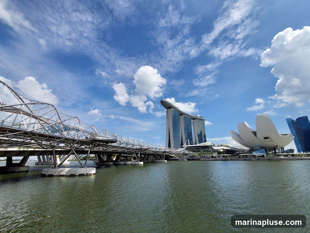 A wide shot of Marina Bay Sands and the Helix Bridge in Singapore under a partly cloudy blue sky. The water in the foreground reflects the sky and buildings.
