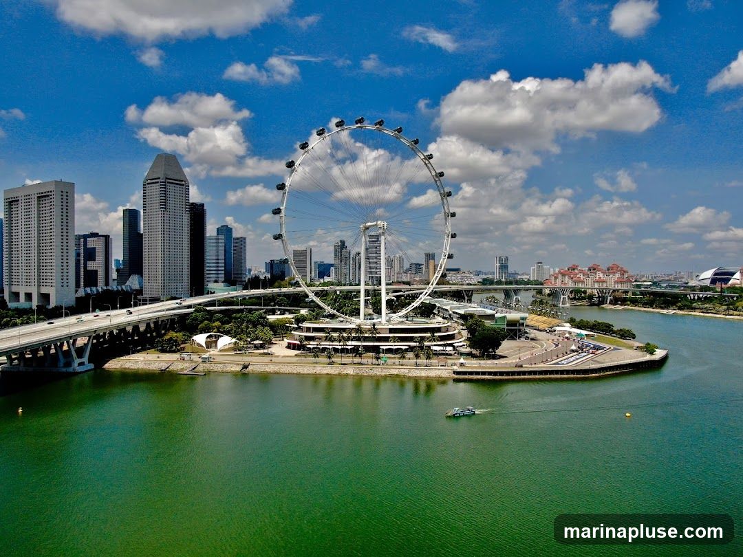 An aerial view of the Singapore skyline featuring the Singapore Flyer, a large Ferris wheel, with modern skyscrapers and a bridge in the background. The green waters of a bay are in the foreground, with a small boat sailing on it. The sky is blue with scattered white clouds.