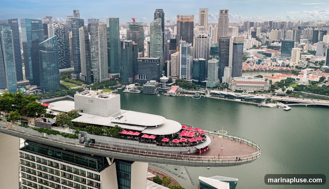 An aerial view of a rooftop bar with numerous red umbrellas, situated on a building that extends outwards over a body of water. The bar area is bustling with patrons. In the background, a dense cityscape of modern skyscrapers is visible under a cloudy sky.