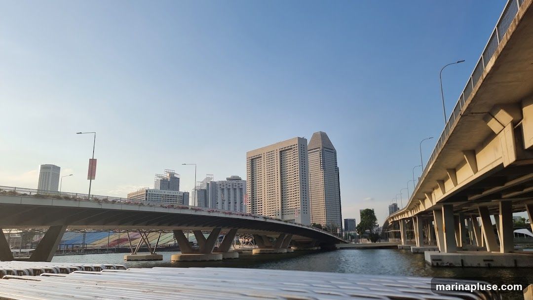 A wide shot of a cityscape featuring multiple bridges spanning a body of water. In the background, several tall buildings rise against a clear blue sky. The foreground shows a portion of a railing or structure, possibly a walkway or seating area, with a metallic, slatted surface. The lighting suggests it is either early morning or late afternoon, casting long shadows and highlighting the architectural elements of the bridges and buildings.