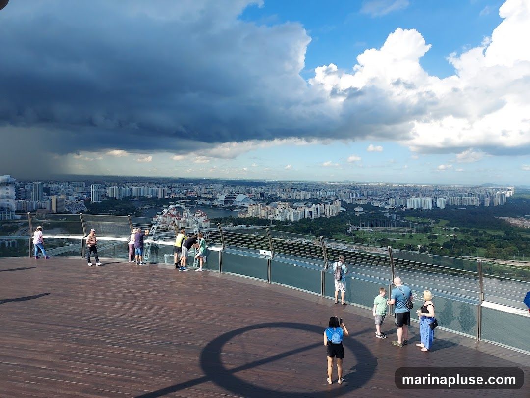 A group of tourists are standing on a wooden observation deck, looking out at a sprawling cityscape under a dramatic sky with dark storm clouds on one side and bright blue sky with fluffy white clouds on the other. The city extends to the horizon, with buildings of various sizes and a Ferris wheel visible in the distance. Some people are taking photos, while others are simply enjoying the panoramic view.