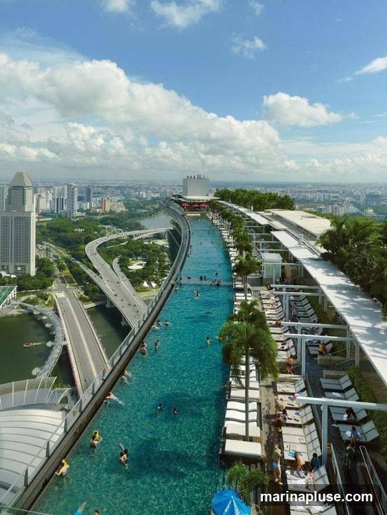 An aerial view of a long, narrow infinity pool situated on a rooftop, overlooking a sprawling cityscape. People are swimming and relaxing in the pool, while lounge chairs are arranged along the poolside. Palm trees add a tropical touch to the scene. In the background, a highway and urban buildings are visible under a partly cloudy sky.
