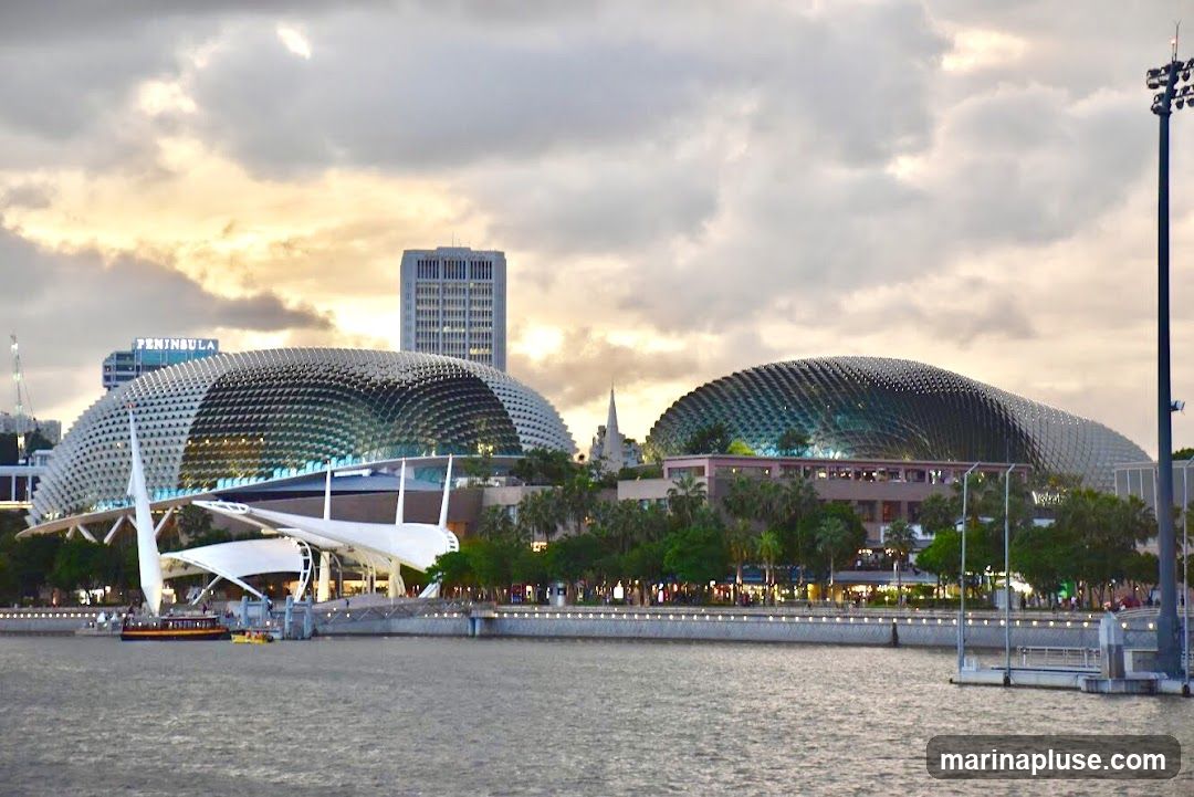 A wide shot of the Marina Bay Sands Theatres in Singapore, with their distinctive durian-shaped domes. The buildings are illuminated against a cloudy sky at dusk. A body of water is in the foreground, with a boat visible on the left. The cityscape and other buildings are visible in the background.