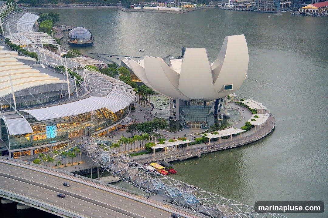An aerial view captures the iconic Marina Bay Sands complex and the ArtScience Museum in Singapore. The ArtScience Museum, with its distinctive lotus-like design, is situated on the waterfront. Adjacent to it is the Marina Bay Sands hotel, a striking architectural landmark. A pedestrian bridge, resembling a coiled helix, connects the two structures. The scene is set against a backdrop of water and other city buildings, with a highway in the foreground.