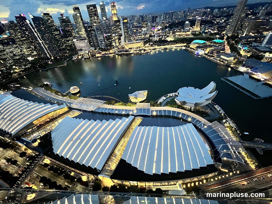 An aerial view of the Singapore skyline at dusk, showcasing the Marina Bay Sands complex and the surrounding cityscape. The iconic lotus-shaped ArtScience Museum is visible, along with the distinctive architecture of the Marina Bay Sands hotel and its rooftop infinity pool. The bay is illuminated, reflecting the lights of the towering skyscrapers in the background. The image captures the vibrant energy and modern development of the city.