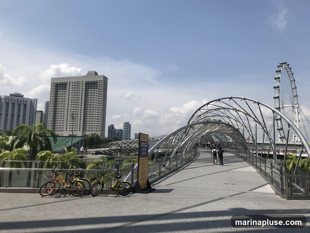 A view of the Helix Bridge in Singapore, with the Singapore Flyer Ferris wheel in the background and a modern city skyline. Several bicycles are parked on the walkway in the foreground. The sky is blue with scattered clouds.