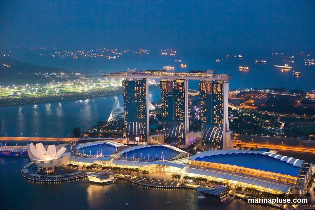 An aerial view of the Marina Bay Sands complex in Singapore at night. The iconic three towers, connected by the SkyPark, are illuminated, as is the lotus-shaped ArtScience Museum and the convention center. The surrounding bay is dotted with ships, and the city skyline glows in the background.