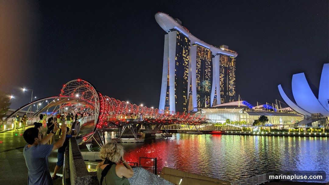 A nighttime panoramic view of the Marina Bay Sands hotel and the Helix Bridge in Singapore. The iconic hotel, with its distinctive boat-shaped top, is illuminated, as are the surrounding buildings and the Helix Bridge, which is lit with red lights. People are visible on the bridge and along the waterfront, taking in the sights. The water reflects the colorful lights of the cityscape.