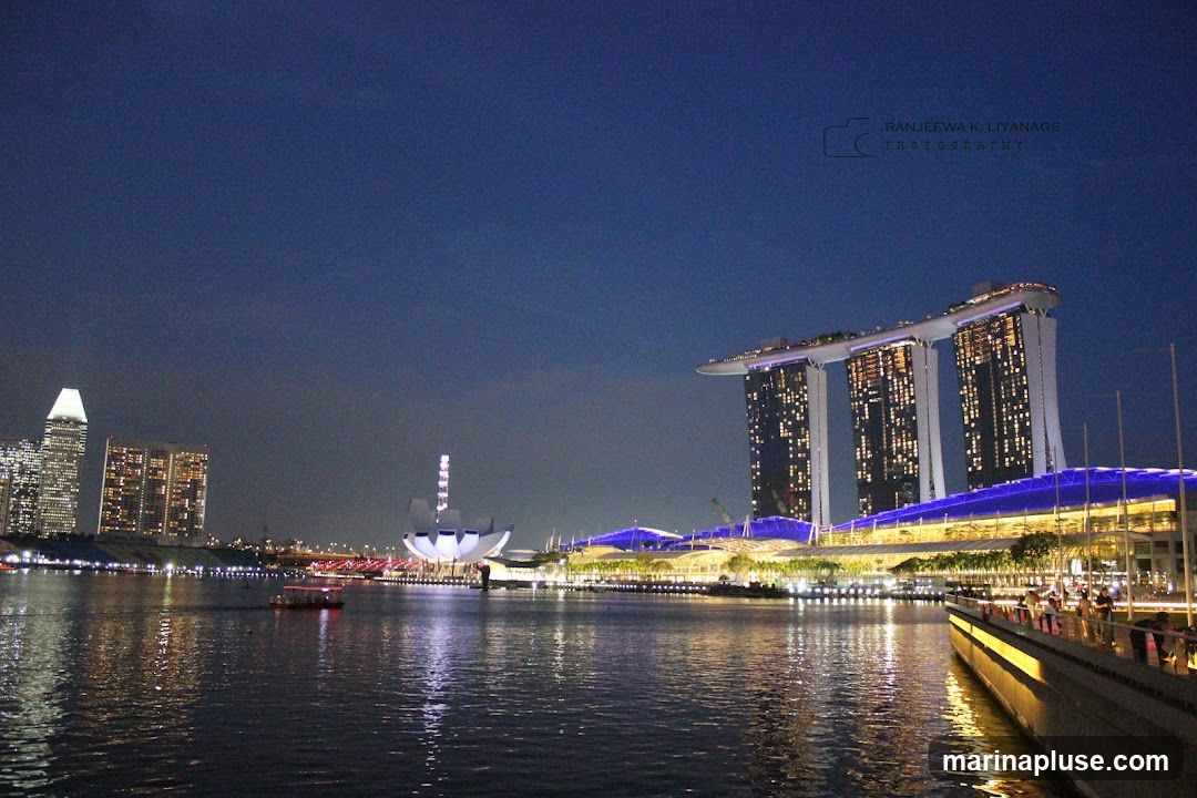 A wide shot of the Marina Bay Sands complex in Singapore at night. The iconic three towers with the sky park on top are illuminated, as is the convention center below. The water in the bay reflects the lights of the buildings and the city skyline in the background. A small boat is visible on the water. A watermark of "RANJEEWA K. LIYANAGE PHOTOGRAPHY" is present in the upper right corner.