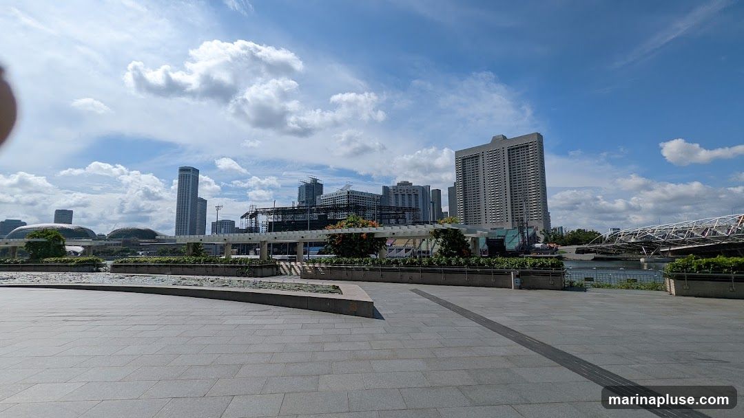 A wide-angle view of a modern cityscape under a partly cloudy blue sky. In the foreground is a paved plaza with planters. In the mid-ground, a white trellis structure with greenery and flowers runs horizontally. Beyond the trellis, the iconic Marina Bay Sands hotel and the Helix Bridge are visible, along with other skyscrapers and buildings. Some construction is also apparent. The overall scene depicts a vibrant urban environment with waterfront views.