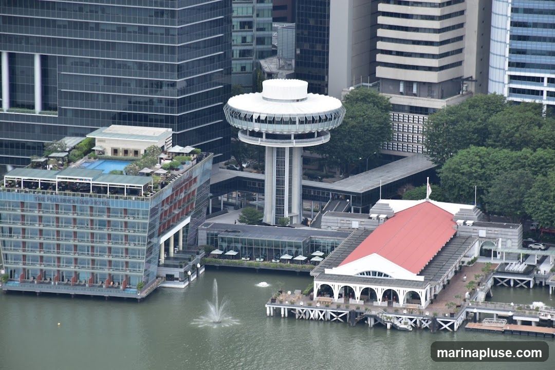An aerial view of several modern buildings situated along a waterfront. On the left is a multi-story hotel with a rooftop pool and seating areas. The hotel's name, 'THE FULLERTON BAY HOTEL', is visible on its facade. In the center, a distinctive tower with a circular observation deck rises above the surrounding structures. To the right, a building with a red roof and arched windows is located on a pier extending into the water. A fountain is active in the water in front of the hotel. The scene is surrounded by lush green trees and other tall city buildings in the background.