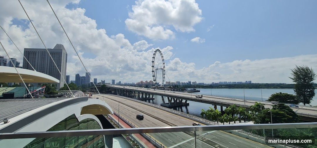 A wide-angle view of a modern cityscape under a partly cloudy sky. In the foreground, a contemporary building with a curved roof and glass facade is visible. Beyond this, a multi-lane highway stretches across a body of water, supported by concrete pillars. A large Ferris wheel stands prominently in the middle ground, with city skyscrapers forming the backdrop. The scene captures a blend of urban infrastructure and recreational landmarks.