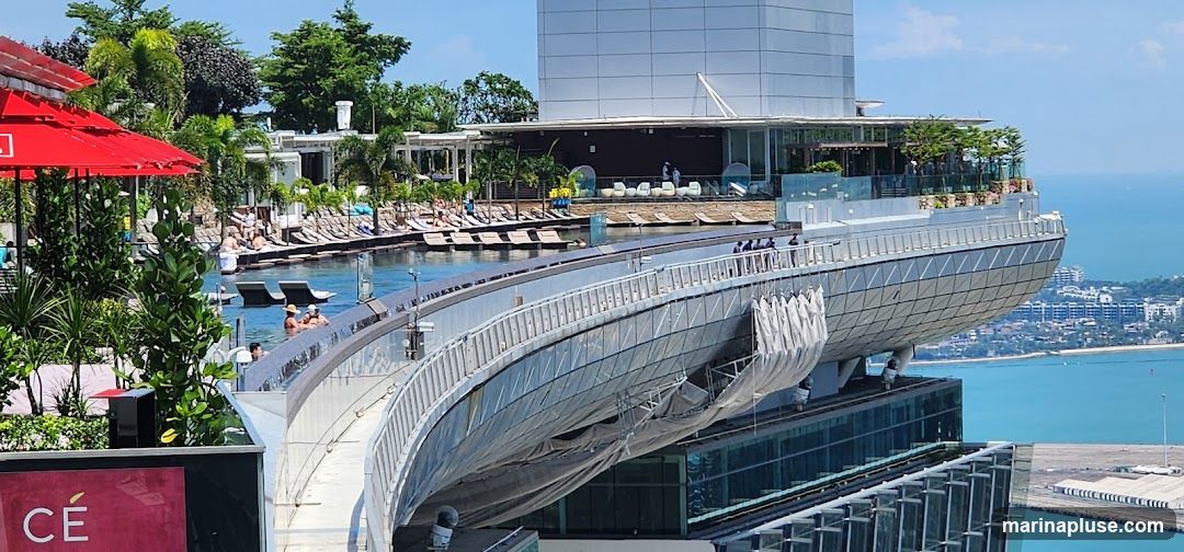 A panoramic view of a luxurious rooftop pool and bar area overlooking a coastal city. The infinity pool is surrounded by lounge chairs and lush greenery. People are seen relaxing by the pool and in the water. The bar area features modern seating and a scenic view of the ocean and cityscape. The architecture is sleek and contemporary, with a prominent curved structure extending from the building.