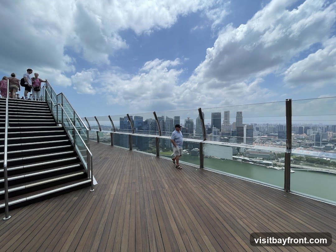 A group of people stand on a wide staircase leading up to an observation deck. The deck has a wooden floor and is bordered by a glass barrier offering a panoramic view of a city skyline and a body of water. A man walks along the deck, looking out at the view. The sky is partly cloudy.